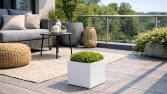 Na nowoczesnym patio znajduje się szara sofa, okrągła wiklinowa otomana, stolik kawowy z imbrykiem, pleciony kosz, rośliny doniczkowe, a na pierwszym planie Donica aluminiowa Alessio 1 wypełniona zielonymi krzewami. Szklana balustrada i drzewa liściaste dopełniają całości.