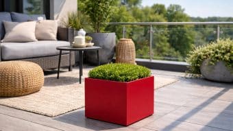 Nowoczesne patio z szarą sofą, neutralnymi poduszkami, okrągłą plecioną otomaną i kwadratową czerwoną Donicą aluminiową Alessio 2 trzymającą zielony krzew. Rośliny zdobią przestrzeń, która wychodzi na zieleń przez szklaną balustradę.