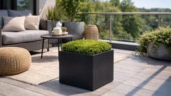 Nowoczesne patio z szarą podłogą wyłożoną kafelkami zawiera Donicę aluminiową Alessio 3 wypełnioną zielonym krzewem, wiklinową otomanę, sofę zewnętrzną z poduszkami, okrągły stolik kawowy i rośliny doniczkowe. Szklana balustrada wychodzi na drzewa przy słonecznej pogodzie.