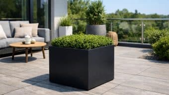 Nowoczesne patio na świeżym powietrzu prezentuje Donica aluminiowa Alessio 4 wypełniona zielonymi krzewami na jasnoszarych płytkach. Za nim, szara sofa z beżowymi poduszkami i okrągły drewniany stolik kawowy siedzą przy szklanych balustradach z widokiem na bujną zieleń.