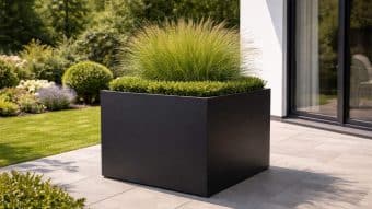 Donica aluminiowa Alessio 5, duża nowoczesna czarna donica, stoi na szarym patio z bujnymi zielonymi krzewami i wysoką trawą ozdobną, podczas gdy wypielęgnowany trawnik i szklane drzwi pojawiają się w słonecznym tle.