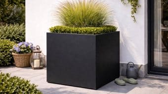 Na szarym patio z trawą ozdobną i przyciętym bukszpanem stoi duża, matowo czarna donica Donica aluminiowa Alessio 6 square. W pobliżu znajdują się zielone kapcie, konewka, niebieskie hortensje w koszyku, latarnia i pnącza przy szklanych drzwiach.