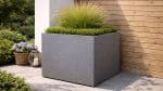 Donica aluminiowa Alessio 7 to nowoczesna, kwadratowa donica w kolorze szarym, w środku której znajdują się starannie przycięte zielone krzewy i wysoka, pierzasta trawa ozdobna. Stoi na wyłożonym kafelkami patio obok drewnianej ściany, w pobliżu latarni i fioletowych kwiatów w koszu.