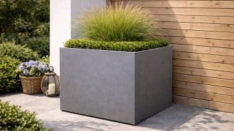 Donica aluminiowa Alessio 7 to nowoczesna, kwadratowa donica w kolorze szarym, w środku której znajdują się starannie przycięte zielone krzewy i wysoka, pierzasta trawa ozdobna. Stoi na wyłożonym kafelkami patio obok drewnianej ściany, w pobliżu latarni i fioletowych kwiatów w koszu.