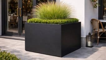 Donica aluminiowa Alessio 8, duża nowoczesna czarna donica, stoi na patio przed szklanymi drzwiami. Umieszczono w niej krzewy bukszpanu w pierścieniu z wysoką trawą ozdobną pośrodku. W pobliżu znajduje się wiklinowe krzesło i lampion ze świecą.