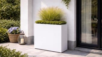 Donica aluminiowa Edoardo 1 stoi na patio przy szklanych drzwiach, wypełnionym zieloną trawą ozdobną i niskimi krzewami bukszpanu. W pobliżu znajduje się wiklinowy kosz z fioletowymi kwiatami i latarnia ze świecą na wyłożonym kafelkami podłożu.