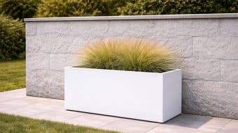 Prostokątna donica Donica aluminiowa Edoardo 2 stoi na szarych płytkach przy jasnoszarej kamiennej ścianie, wypełnionej gęstą trawą ozdobną. Za ścianą widoczna jest zielona trawa i krzewy.