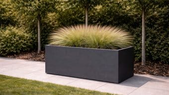 Donica aluminiowa Edoardo 5, prostokątna aluminiowa donica malowana proszkowo na czarno, mieści pierzastą trawę ozdobną. Stoi na szarej kostce brukowej przy zielonym trawniku, otoczona liściastymi drzewami i krzewami w ogrodzie krajobrazowym.