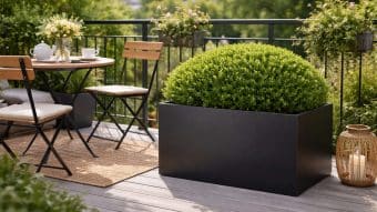 Przytulny balkon ma okrągły stół z kwiatami, zestaw do herbaty i dwa składane krzesła. Donica aluminiowa Fabio Mobile podtrzymuje okrągły zielony krzew. Latarnie i rośliny doniczkowe dodają uroku wśród bujnej zieleni na balustradzie.