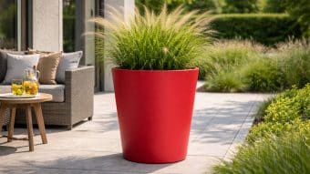 Donica aluminiowa Silvestro 2, wypełniona wysoką zieloną trawą ozdobną, stoi na nasłonecznionym patio obok wiklinowego fotela z beżowymi poduszkami i okrągłego drewnianego stołu z lemoniadą i szklankami. Całości dopełnia bujna zieleń.