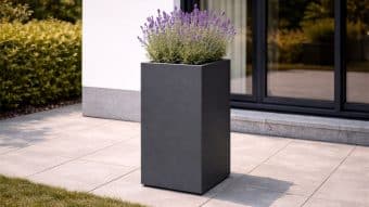 Wysoka, wąska Donica aluminiowa Vittorio 1 w kolorze matowej czerni z kwitnącą lawendą stoi na wyłożonym beżowymi kafelkami patio w pobliżu nowoczesnego budynku z ciemnymi szklanymi drzwiami i białymi ścianami, z zielonym żywopłotem i trawą widoczną w tle.