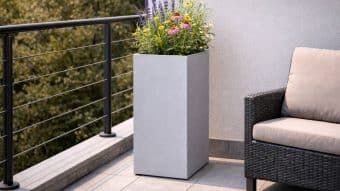 Donica aluminiowa Vittorio 2, wypełniona kolorowymi kwiatami, stoi na wyłożonym kafelkami balkonie obok ciemnego wiklinowego krzesła z beżowymi poduszkami. Metalowe barierki ograniczają przestrzeń, a w tle widać drzewa.