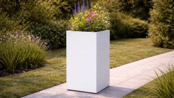 Donica aluminiowa Vittorio 3, wysoka prostokątna donica aluminiowa, stoi na kamiennej ścieżce w ogrodzie wypełnionym kolorowymi polnymi kwiatami pośród zielonych krzewów. Światło słoneczne podkreśla jej czyste linie i nowoczesny design.