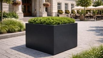 Donica aluminiowa Alessio 10 z gęstymi zielonymi krzewami znajduje się na brukowanym placu przed eleganckim budynkiem z kolumnami, przyciętymi krzewami, wiszącymi koszami kwiatowymi i zacienionymi miejscami do siedzenia na zewnątrz.