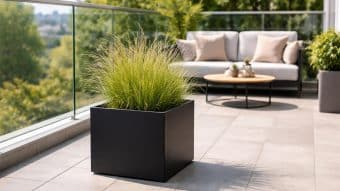 Nowoczesny balkon ze szklaną balustradą prezentuje Donica aluminiowa Alessio 11, idealny jako donice do biura i wypełniony wysoką zieloną trawą. Beżowa sofa z poduszkami i okrągły drewniany stolik kawowy zapewniają komfort, a w tle widać bujną zieleń.