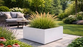 W nowoczesnym patio znajduje się Donica aluminiowa Alessio 12 z trawą ozdobną, otoczona wiklinową sofą z szarymi poduszkami, okrągłym stołem ze świecą, kwitnącymi kwiatami i bujną zielenią w jasnym świetle dziennym.