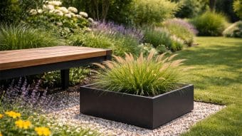 Nowoczesna scena ogrodowa z drewnianą ławką z czarną metalową ramą na żwirze. Obok stoi Donica aluminiowa Alessio 14 wypełniona trawą ozdobną, otoczona kwitnącymi kwiatami i bujną zielenią w jasnym świetle słonecznym.