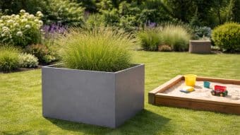 Duża, szara, kwadratowa donica Donica aluminiowa Alessio 15 z wysoką trawą ozdobną stoi na starannie skoszonym trawniku obok drewnianej piaskownicy z piaskiem, żółtym wiaderkiem i czerwoną wywrotką. Bujne krzewy i kwitnące rośliny tworzą tło.