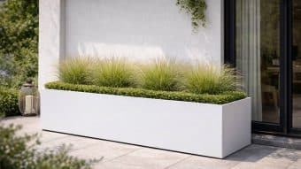 Donica aluminiowa Alessio 9, nowoczesna prostokątna biała donica, stoi na wyłożonym kafelkami patio przy szklanych drzwiach z zasłoną. Jest wypełniona zielonymi krzewami i wysokimi trawami, podkreślonymi przez dekoracyjną latarnię w pobliżu.