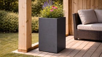 Donica aluminiowa Vittorio 4, wysoka i ciemnoszara, jest wypełniona kolorowymi kwiatami na drewnianym patio obok wiklinowej sofy zewnętrznej z beżowymi poduszkami. Światło słoneczne wpada do środka, a w tle widać zielone liście.