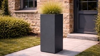 Nowoczesna scena na zewnątrz prezentuje wysoką, prostokątną Donicę aluminiową Vittorio 5 z trawą ozdobną. Donica spoczywa na szarych kamiennych płytkach przy budynku z kamienną ścianą, oknem, ciemnymi drzwiami i bujnymi zielonymi krzewami i trawą w pobliżu.