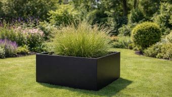 Duża Donica aluminiowa Alessio 13, wypełniona wysoką trawą ozdobną, stoi na starannie skoszonym trawniku otoczonym zielonymi krzewami, kwitnącymi kwiatami i drzewami w tętniącym życiem, oświetlonym słońcem ogrodzie.