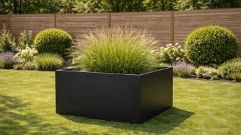 Duża, prostokątna Donica aluminiowa Alessio 17 wypełniona wysoką trawą ozdobną stoi na wypielęgnowanym trawniku. W tle znajdują się zadbane krzewy, kwitnące rośliny i drewniany płot, tworząc nowoczesną scenę ogrodową skąpaną w słońcu.