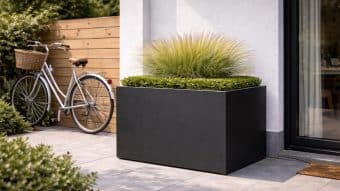 W nowoczesnym patio znajduje się Donica aluminiowa Edoardo 10, wypełniona ozdobną trawą i zielonymi krzewami, w pobliżu białej ściany i szklanych drzwi. W tle zabytkowy rower opiera się o drewniany płot pośród krzewów i płytek.