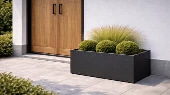 Donica aluminiowa Edoardo 11 w nowoczesnej czerni prezentuje trzy okrągłe, przycięte zielone krzewy i wysoką trawę ozdobną na wyłożonym kafelkami patio w pobliżu białej ściany i podwójnych drzwi z jasnego drewna z czarnymi klamkami, otoczonych niską zielenią.