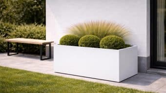 Donica aluminiowa Edoardo 12, w nowoczesnym białym kolorze, prezentuje trzy krzewy o okrągłym kształcie i trawę ozdobną na szarym patio wyłożonym kafelkami przy białym budynku. Obok stoi drewniana ławka z czarnymi nogami, zwrócona w stronę zielonego trawnika i bujnych krzewów.