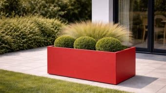 Donica aluminiowa Edoardo 13, nowoczesna czerwona prostokątna donica, prezentuje trzy okrągłe zielone krzewy i wysoką delikatną trawę na wyłożonym kafelkami patio przy szklanych drzwiach, z żywopłotami i trawnikiem w tle.