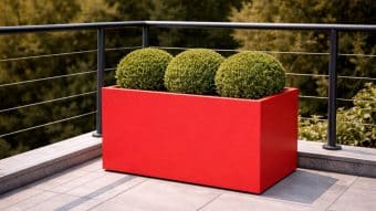 Donica aluminiowa Edoardo 8 w jaskrawoczerwonym kolorze trzyma trzy starannie przycięte okrągłe zielone krzewy na nowoczesnym patio z metalową balustradą, szarymi płytkami i drzewami w tle.