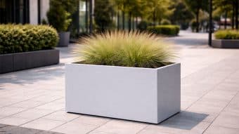 Donica aluminiowa Edoardo 9, duża prostokątna donica z wysoką trawą ozdobną, jest umieszczona na wybrukowanym chodniku w nowoczesnym otoczeniu miejskim, otoczonym zielenią i drzewami.