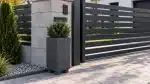 Czarna wysoka donica ogrodowa z zielonym krzewem przy nowoczesnej bramie wjazdowej.