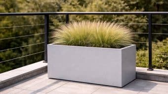 Duża, szara Donica aluminiowa Edoardo 14 obsypana wysoką, pierzastą trawą ozdobną na wyłożonym kafelkami balkonie z czarną metalową balustradą. Zielone drzewa liściaste tworzą delikatnie rozmyte tło za balustradą.