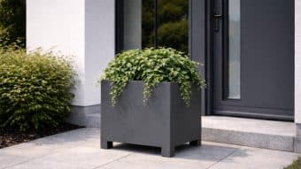 Duża, kwadratowa, ciemnoszara Donica aluminiowa Apollo 1 stoi na wyłożonym kafelkami patio w pobliżu nowoczesnych szklanych drzwi, z doniczką białą na nóżkach i zielonym krzewem widocznym w tle przy wejściu do współczesnego domu.