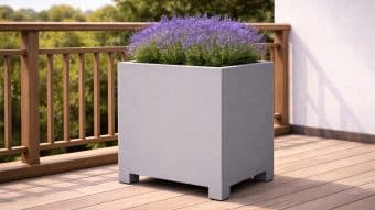 Donica aluminiowa Apollo 2 w kolorze jasnoszarym, mierząca 60 cm i wypełniona fioletową lawendą, stoi na drewnianym tarasie obok drewnianej balustrady. Światło słoneczne oświetla zielone drzewa i białą stiukową ścianę w tle.