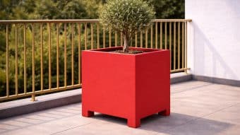 Małe drzewko rośnie w Donica aluminiowa Apollo 3 na słonecznym balkonie z beżowymi płytkami, złotą metalową balustradą, zielonymi drzewami w tle i białą ścianą po prawej stronie.