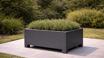 Donica aluminiowa Apollo 5, duża i prostokątna w kolorze czarnym na nóżkach, stoi na szarych płytkach zewnętrznych z gęstymi zielonymi roślinami. Trawa i bujne krzewy w tle tworzą spokojną atmosferę ogrodu.