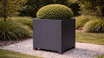 Duża, nowoczesna Donica aluminiowa Apollo 11 Mobile w matowej czerni stoi przy wejściu, trzymając starannie przycięty okrągły zielony krzew, ustawiony na żwirowej ścieżce wśród wypielęgnowanych trawników i bujnej zieleni ogrodowej.