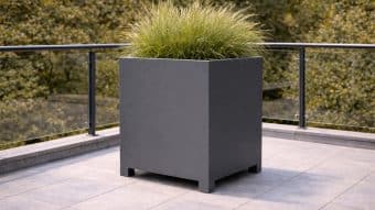 Duża, kwadratowa Donica aluminiowa Apollo 11 wypełniona wysoką trawą ozdobną stoi na wyłożonym płytkami tarasie ze szklanymi balustradami. Liściaste zielone drzewa rozmywają się w tle, tworząc jasną i nowoczesną scenę na zewnątrz.