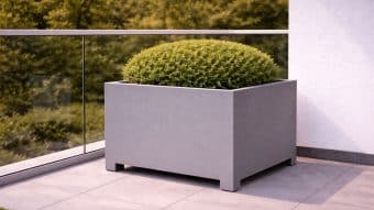 Okrągły, starannie przycięty zielony krzew stoi w dużej, nowoczesnej Donica aluminiowa Apollo 13 Mobile na wyłożonym kafelkami balkonie. Szklana balustrada i bujne drzewa znajdują się w tle, z częścią białej ściany po prawej stronie.