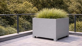 Duża szara Donica aluminiowa Apollo 13, obsadzona wysoką zieloną trawą ozdobną, stoi na wyłożonym kafelkami tarasie. Szklane i metalowe balustrady otaczają ten obszar, a bujne drzewa w tle podkreślają nowoczesną scenerię zewnętrzną.