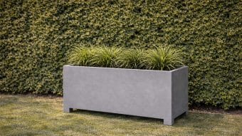 Donica aluminiowa Apollo 14 Mobile stoi na trawniku przed zadbanym zielonym żywopłotem, a jej prostokątny kształt wypełniają równomiernie rozmieszczone kępki zdrowej, kolczastej zielonej trawy.