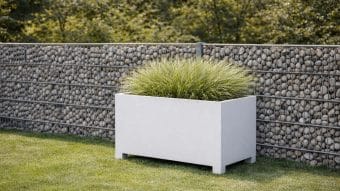 Donica aluminiowa Apollo 14 na nogach, wypełniona trawą ozdobną, stoi na zielonym trawniku przed ogrodzeniem z metalowych klatek z szarymi kamieniami, z liściastymi drzewami tworzącymi bujne tło.