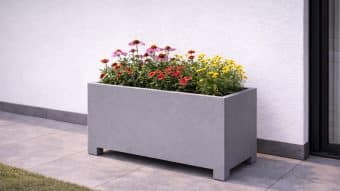 Donica aluminiowa Apollo 15 Mobile stoi na wyłożonym kafelkami patio przy białej ścianie, wypełniona kwitnącymi czerwonymi, żółtymi i różowymi kwiatami. Nieznacznie uniesiona nad ziemią, donica stoi obok skrawka trawy.