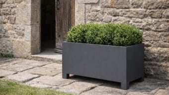 Donica aluminiowa Apollo 15 w kolorze ciemnoszarym, wypełniona starannie przyciętymi zielonymi krzewami, stoi na kamiennym bruku przy rustykalnej kamiennej ścianie i częściowo otwartych drewnianych drzwiach, przywołując naturalną atmosferę na zewnątrz z trawą na pierwszym planie.