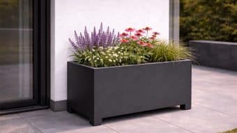 Prostokątna, ciemnoszara donica aluminiowa Apollo 16 Mobile (60 cm wysokości) stoi na wyłożonym kafelkami patio przy szklanych drzwiach i białej ścianie, wypełniona fioletową szałwią, różowymi jeżówkami, małymi białymi kwiatami i trawami ozdobnymi. W tle widać zielone krzewy.