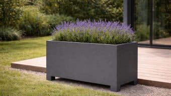 Donica aluminiowa Apollo 16 w kolorze białym, wypełniona kwitnącą lawendą, stoi na żwirze obok drewnianego tarasu, z bujnym zielonym trawnikiem i gęstymi drzewami w tle, tworząc spokojną atmosferę podwórka w słoneczny dzień.