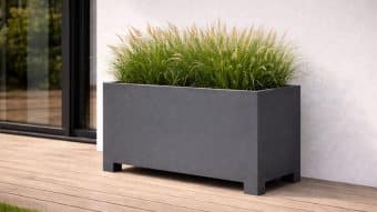 Donica aluminiowa Apollo 17 w prostokątnym szarym kolorze utrzymuje wysokie, bujne trawy na jasnym drewnianym tarasie przy białej ścianie i szklanych drzwiach, wprowadzając miękką teksturę do minimalistycznej przestrzeni zewnętrznej.